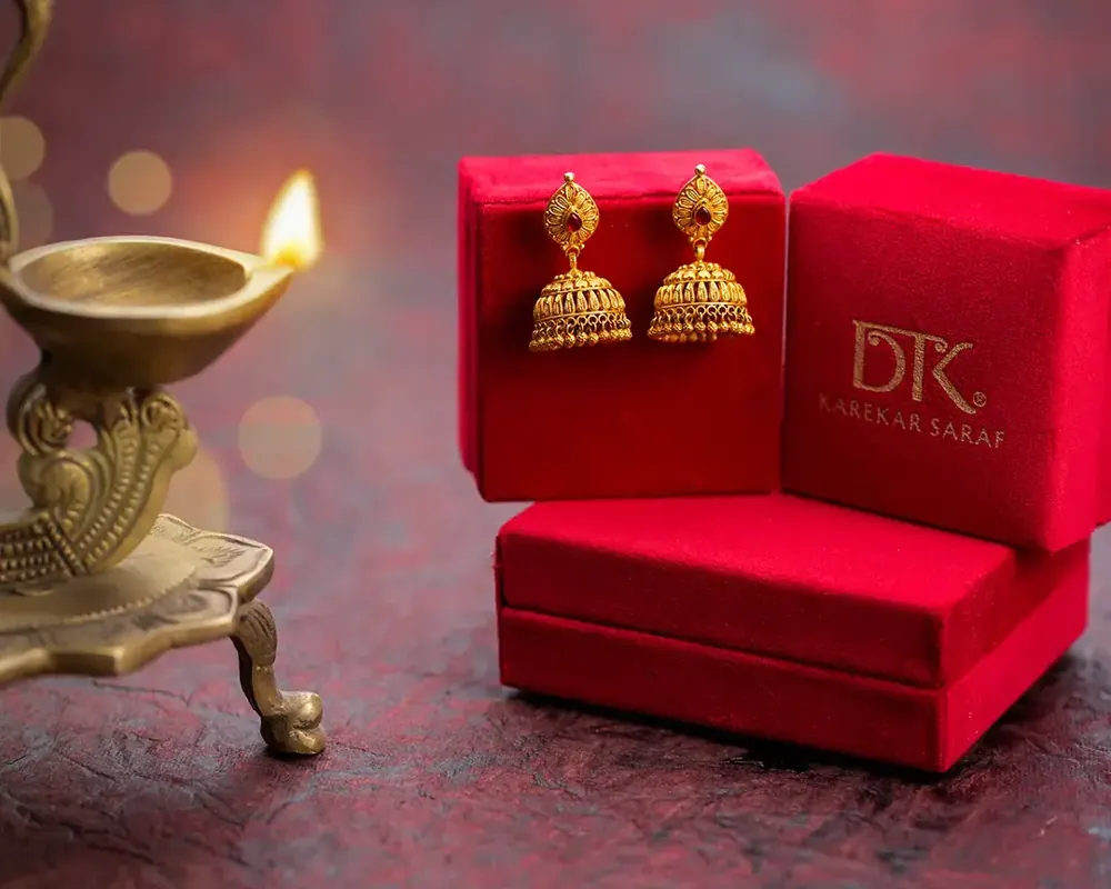 Gold jhumka earrings in a red box beside a diya